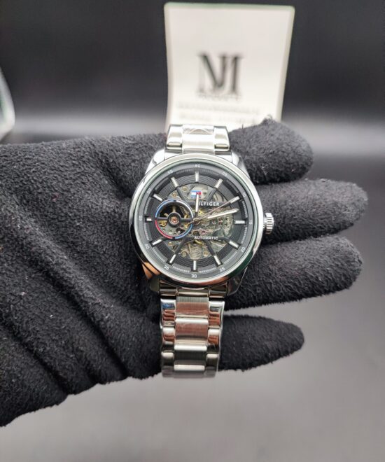 Tommy Hilfiger style men's automatic skeleton watch featuring a polished stainless steel case and link bracelet, a dark grey outer dial ring, and an exposed mechanical movement with a multi-colored subdial accent ring held in a black gloved hand.