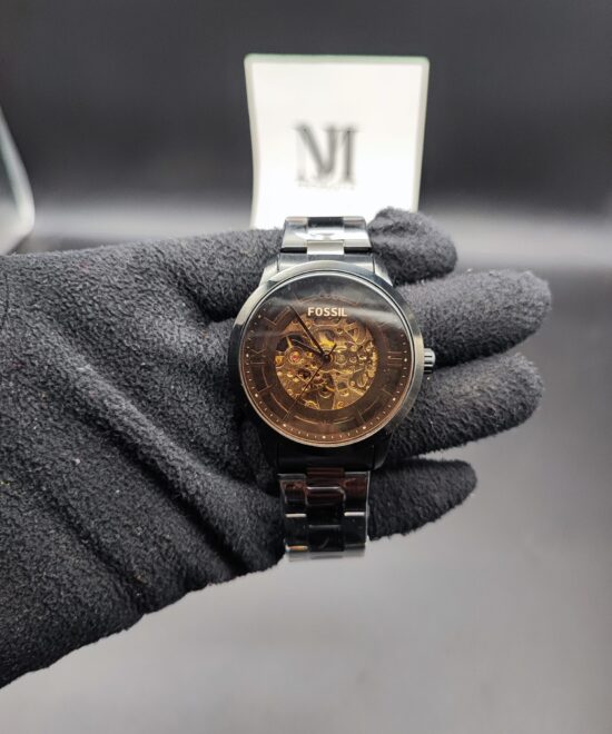 Fossil style men's automatic skeleton watch featuring a polished black case and bracelet, a smoked transparent dial ring, and an exposed gold-tone mechanical movement held in a black gloved hand.