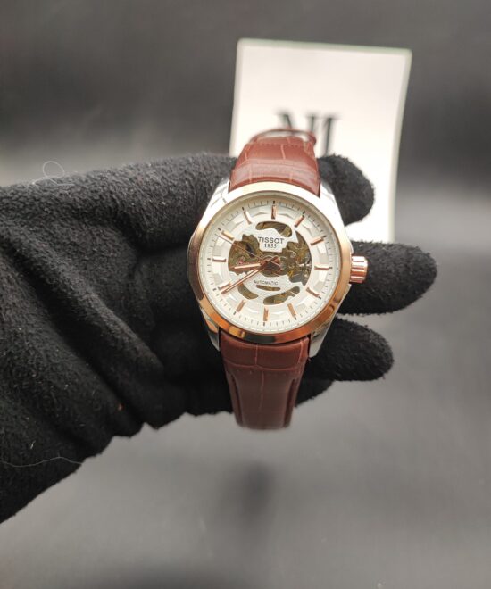 Tissot style open-heart skeleton automatic watch with a rose gold bezel, white outer dial ring, and a brown alligator-texture leather strap held in a black gloved hand.