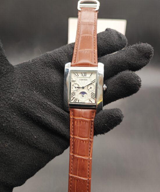 Cartier style rectangular tank dress watch featuring a polished silver case, white guilloché dial with Roman numerals, day-date indicators, moonphase subdial, and a brown alligator-textured leather strap held in a black gloved hand.