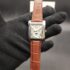Cartier style rectangular tank dress watch featuring a polished silver case, white guilloché dial with Roman numerals, day-date indicators, moonphase subdial, and a brown alligator-textured leather strap held in a black gloved hand.