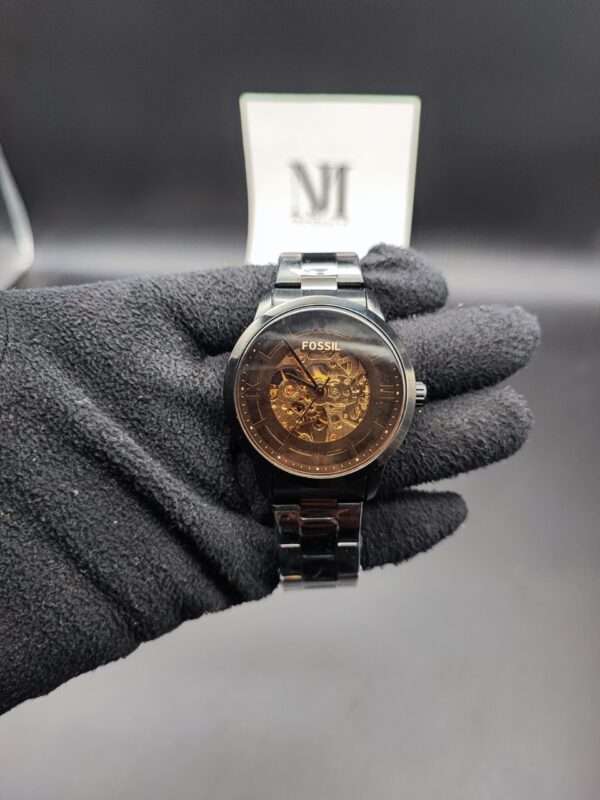 Fossil style men's automatic skeleton watch featuring a polished black case and bracelet, a smoked transparent dial ring, and an exposed gold-tone mechanical movement held in a black gloved hand.