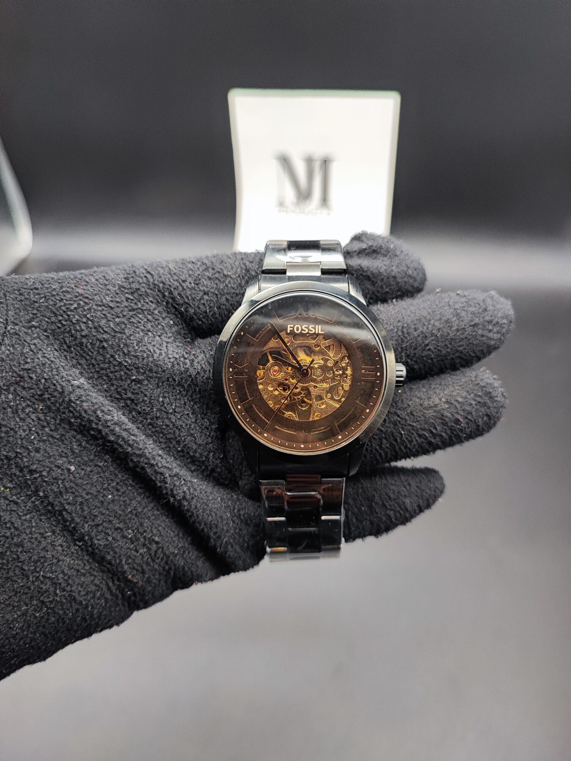 Fossil style men's automatic skeleton watch featuring a polished black case and bracelet, a smoked transparent dial ring, and an exposed gold-tone mechanical movement held in a black gloved hand.