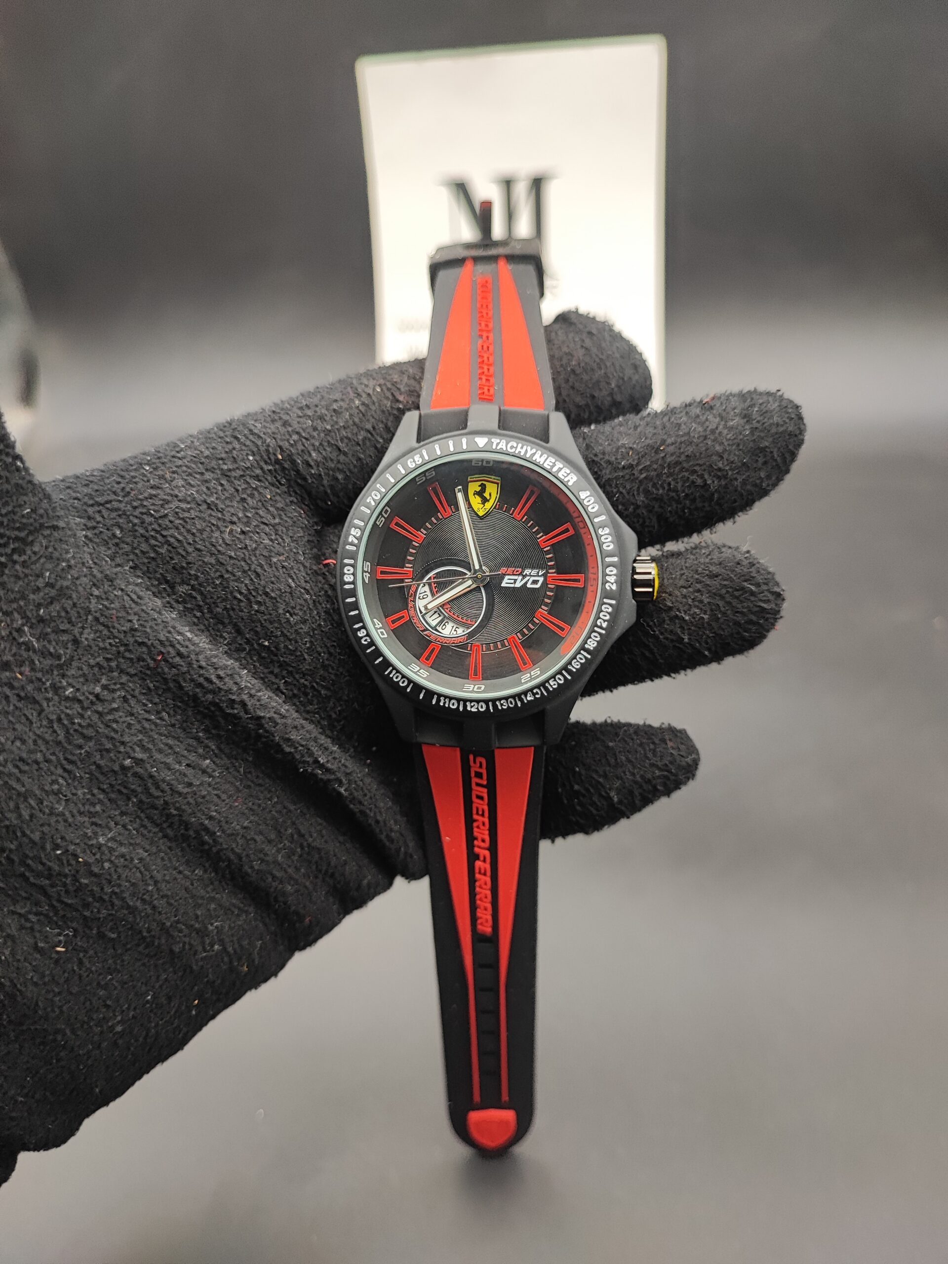 Scuderia Ferrari Red Rev Evo style men's sports watch featuring a matte black case, black honeycomb dial with red index hour markers, yellow racing shield emblem at 12 o'clock, and a red-striped black silicone strap held in a black gloved hand.