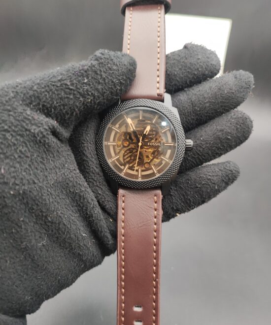 : Fossil style men's automatic skeleton watch featuring a black knurled textured bezel, exposed gold-tone mechanical movement, and a smooth brown leather strap with contrast stitching held in a black gloved hand.