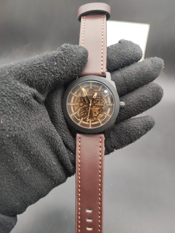 : Fossil style men's automatic skeleton watch featuring a black knurled textured bezel, exposed gold-tone mechanical movement, and a smooth brown leather strap with contrast stitching held in a black gloved hand.