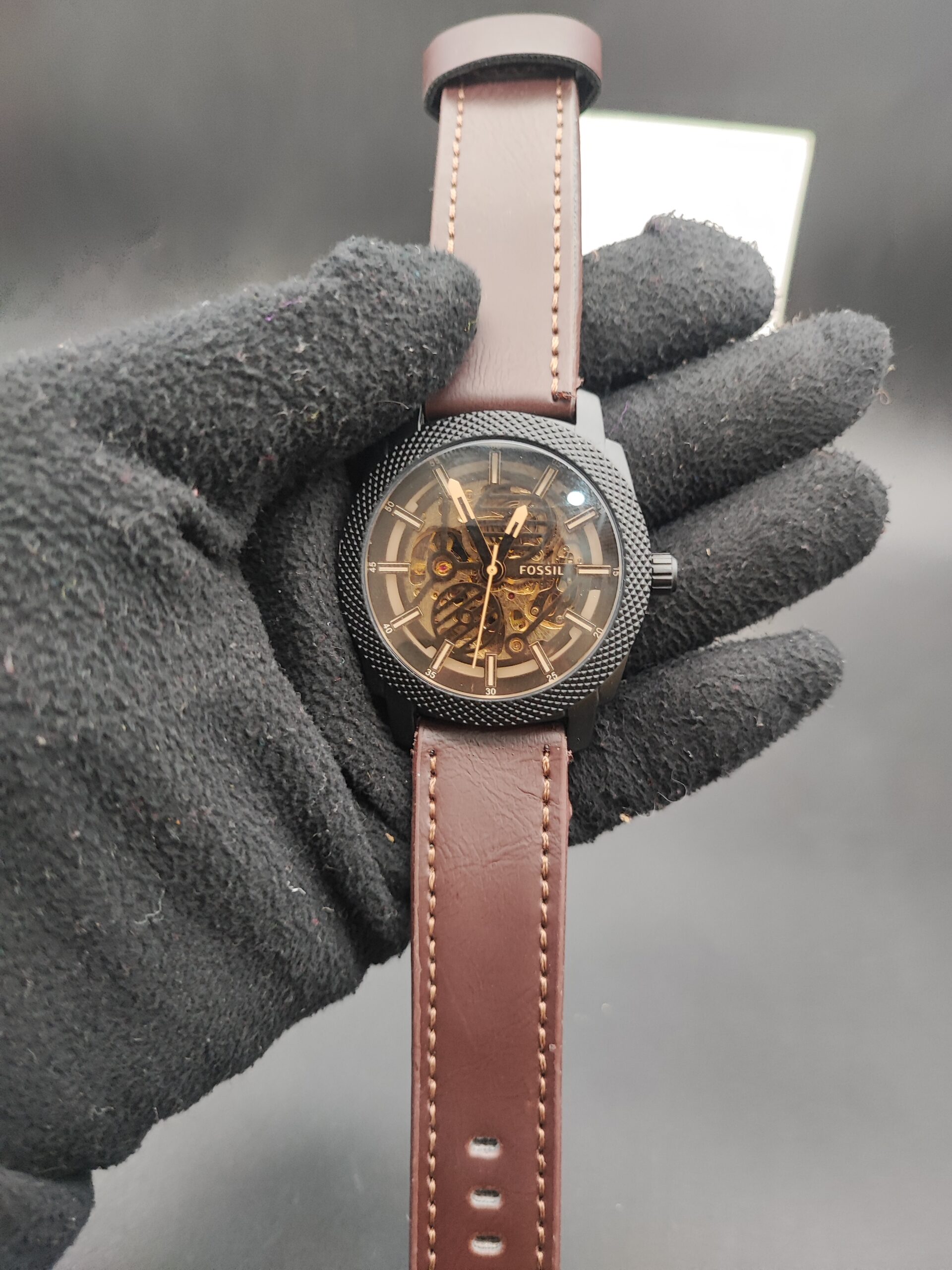 : Fossil style men's automatic skeleton watch featuring a black knurled textured bezel, exposed gold-tone mechanical movement, and a smooth brown leather strap with contrast stitching held in a black gloved hand.
