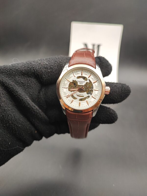 Tissot style open-heart skeleton automatic watch with a rose gold bezel, white outer dial ring, and a brown alligator-texture leather strap held in a black gloved hand.