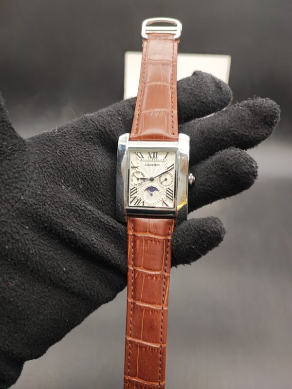 Cartier style rectangular tank dress watch featuring a polished silver case, white guilloché dial with Roman numerals, day-date indicators, moonphase subdial, and a brown alligator-textured leather strap held in a black gloved hand.