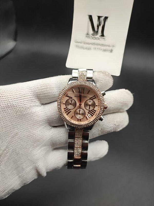 ichael Kors style women's dual-tone chronograph watch featuring a crystal-studded rhinestone bezel, rose gold sunray dial with Roman numerals, and a silver bracelet with diamond-encrusted center links held in a white gloved hand.