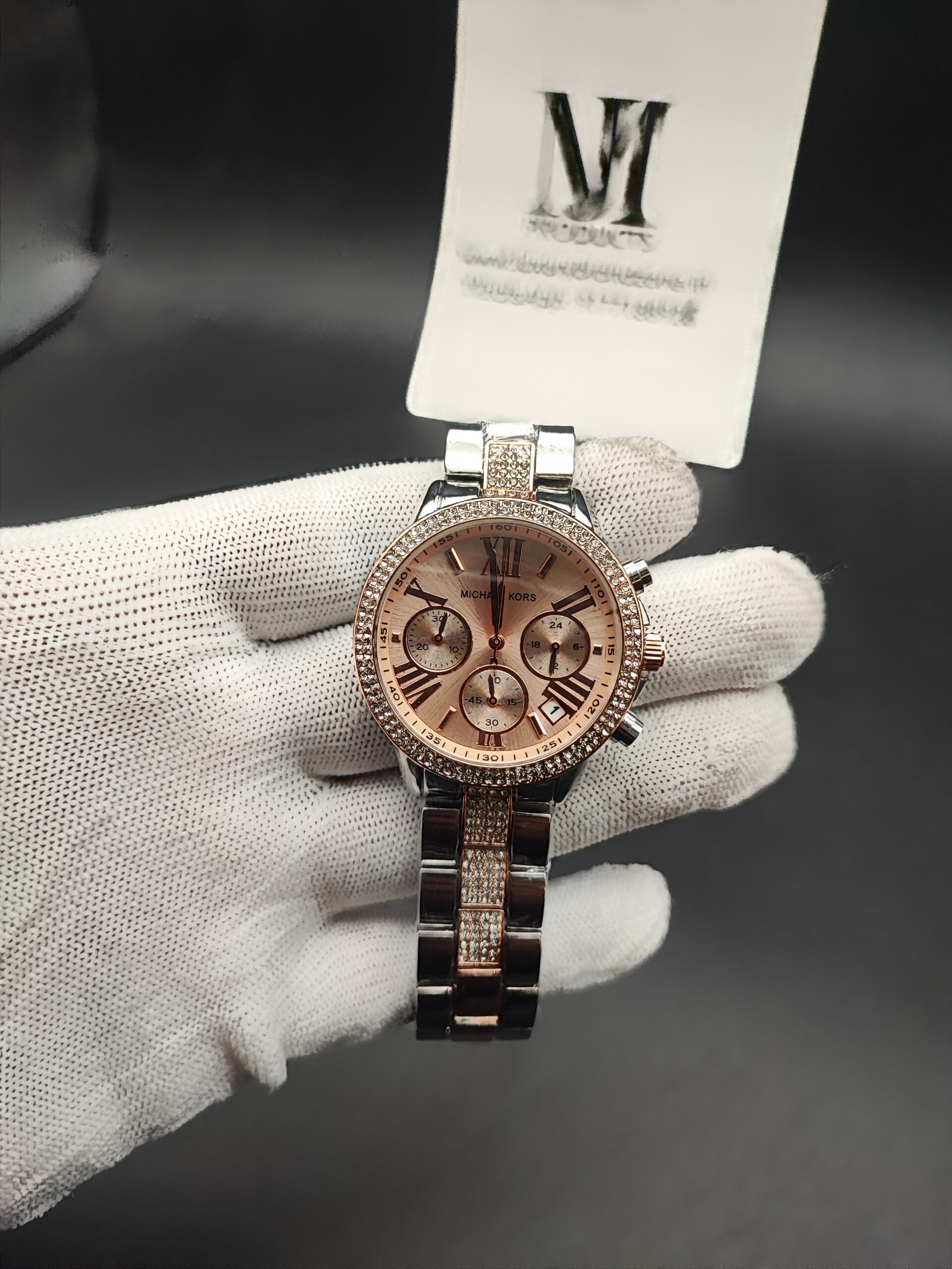 ichael Kors style women's dual-tone chronograph watch featuring a crystal-studded rhinestone bezel, rose gold sunray dial with Roman numerals, and a silver bracelet with diamond-encrusted center links held in a white gloved hand.