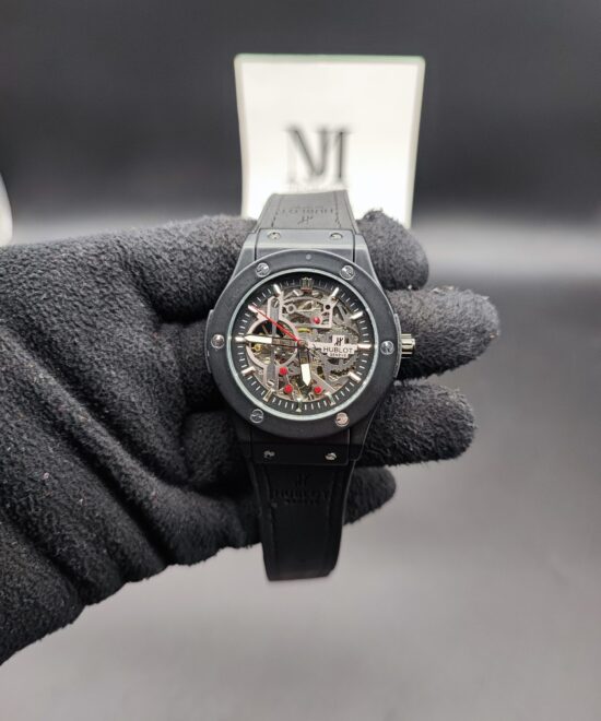 Hublot style Classic Fusion men's skeleton automatic watch featuring a matte black bezel with exposed silver screws, a fully open-worked mechanical skeleton dial with silver gears and a red seconds hand, attached to a black rubber strap held in a black gloved hand.