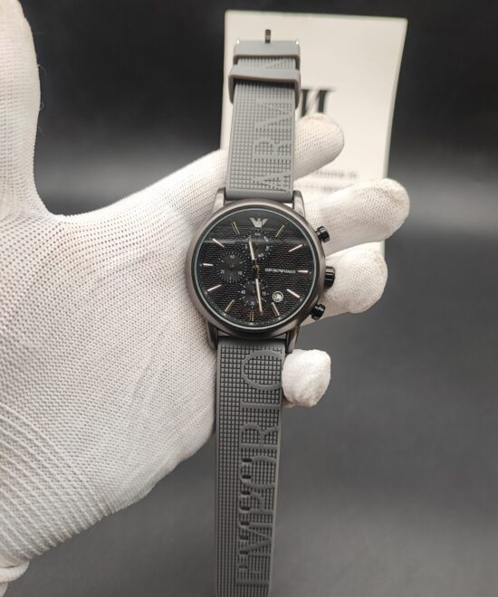 Emporio Armani style men's watch featuring a gunmetal grey case, a black horizontal-paved dial with triple subdials, a silver eagle logo at 12 o'clock, and a textured grey rubber strap with embossed lettering held in a white gloved hand.