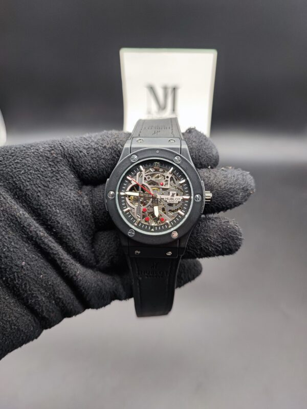Hublot style Classic Fusion men's skeleton automatic watch featuring a matte black bezel with exposed silver screws, a fully open-worked mechanical skeleton dial with silver gears and a red seconds hand, attached to a black rubber strap held in a black gloved hand.