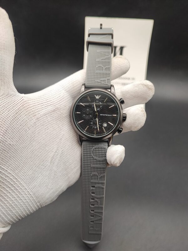 Emporio Armani style men's watch featuring a gunmetal grey case, a black horizontal-paved dial with triple subdials, a silver eagle logo at 12 o'clock, and a textured grey rubber strap with embossed lettering held in a white gloved hand.