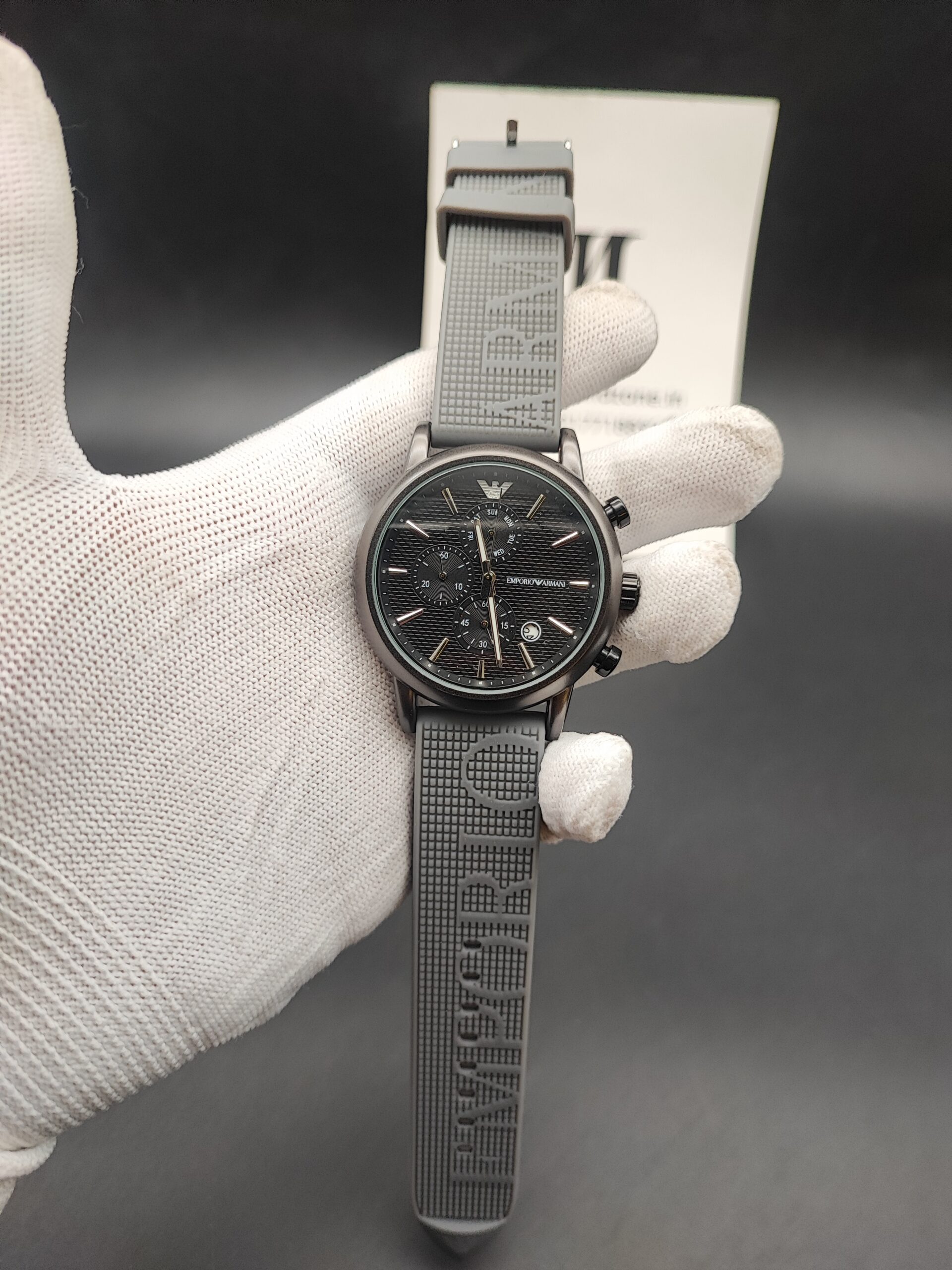 Emporio Armani style men's watch featuring a gunmetal grey case, a black horizontal-paved dial with triple subdials, a silver eagle logo at 12 o'clock, and a textured grey rubber strap with embossed lettering held in a white gloved hand.